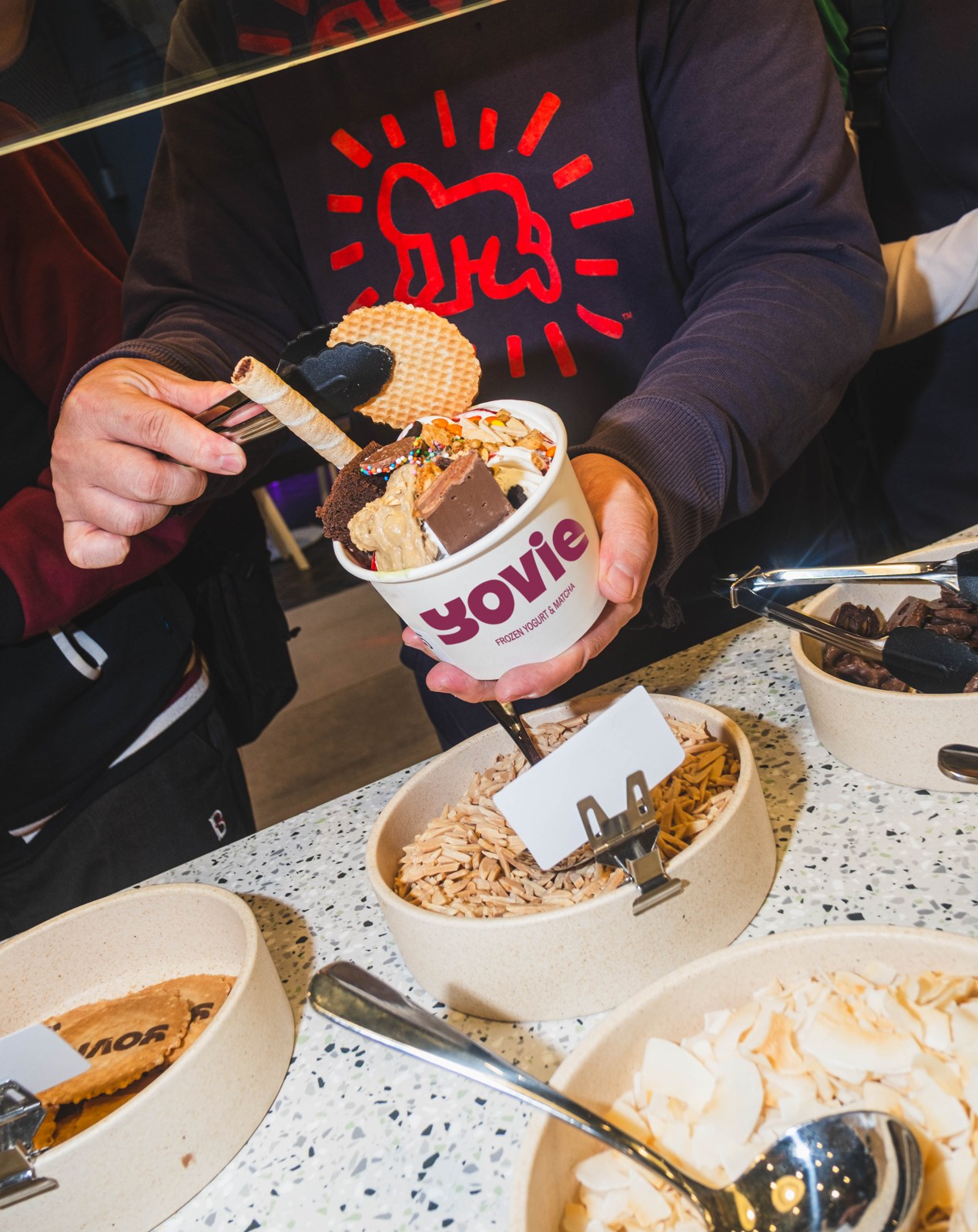 A customer adding toppings to a YOVIE Frozen Yogurt & Matcha cup at the self-serve topping bar