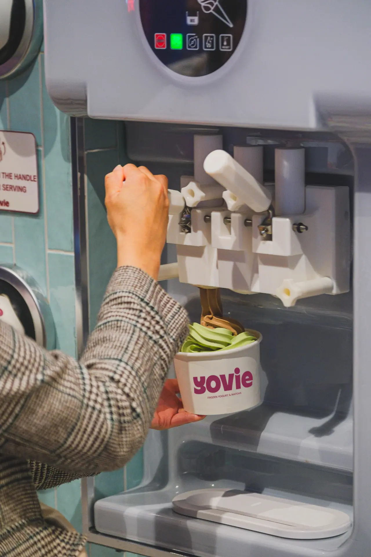 A customer pulling the dispenser handle to serve matcha frozen yogurt into a YOVIE cup